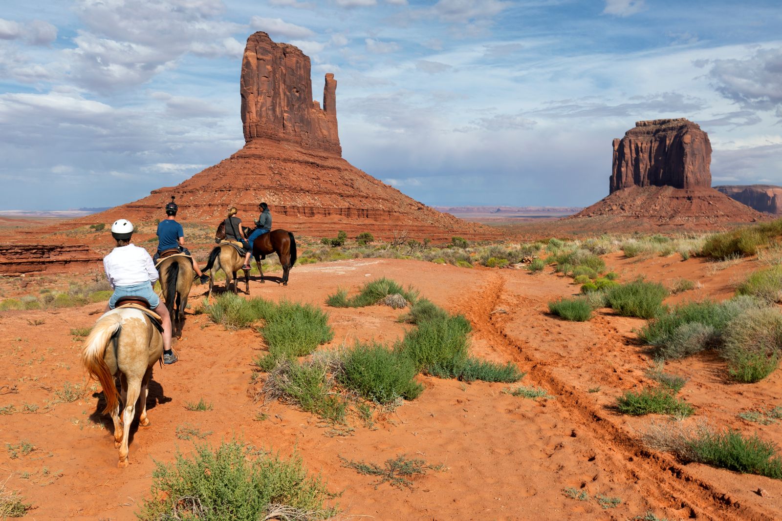 Riders on horseback crossing red sand toward a tall butte